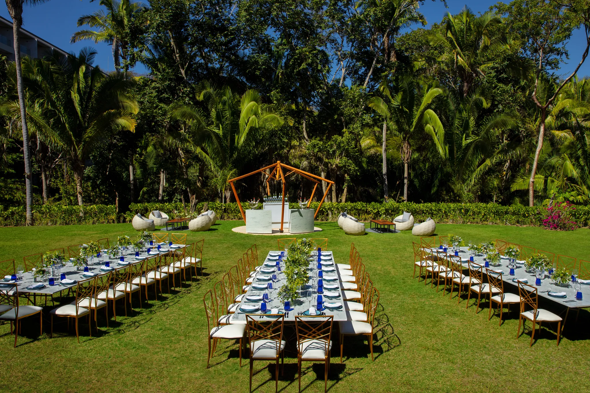 drbmigrpgardenbanquet2acb be all inclusive Outdoor gala setup on a green lawn with long white-clothed tables, gold chairs, and blue napkins, surrounded by tropical palm trees and greenery.