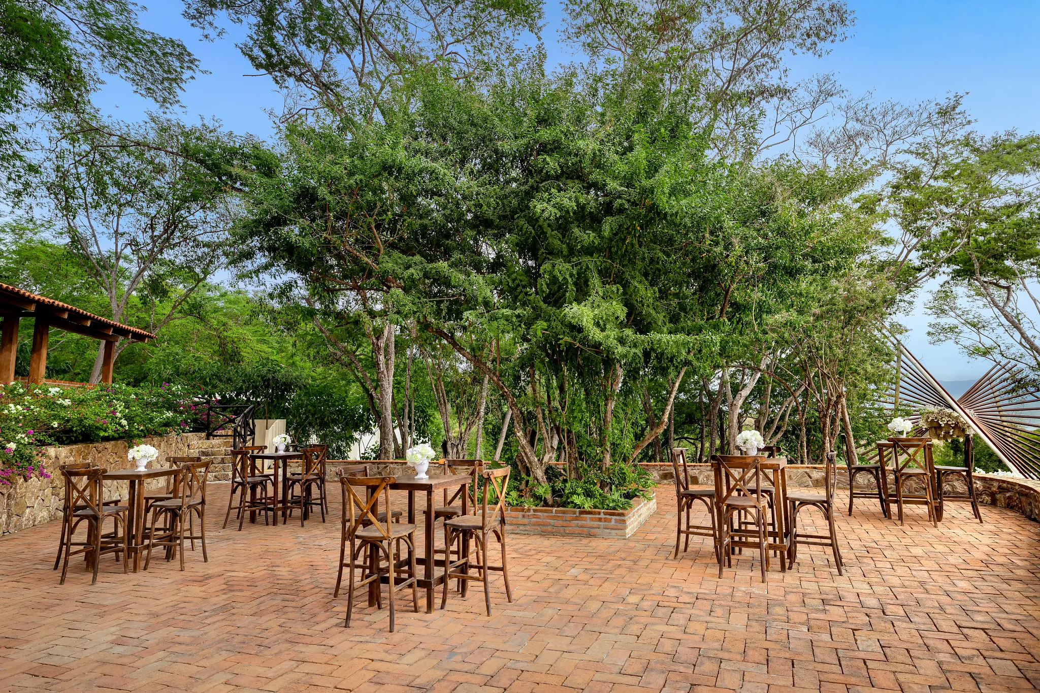 drbmiresthenestafc be all inclusive Outdoor cafe patio with many wooden tables and chairs on brick paving, surrounded by green trees and flowers.