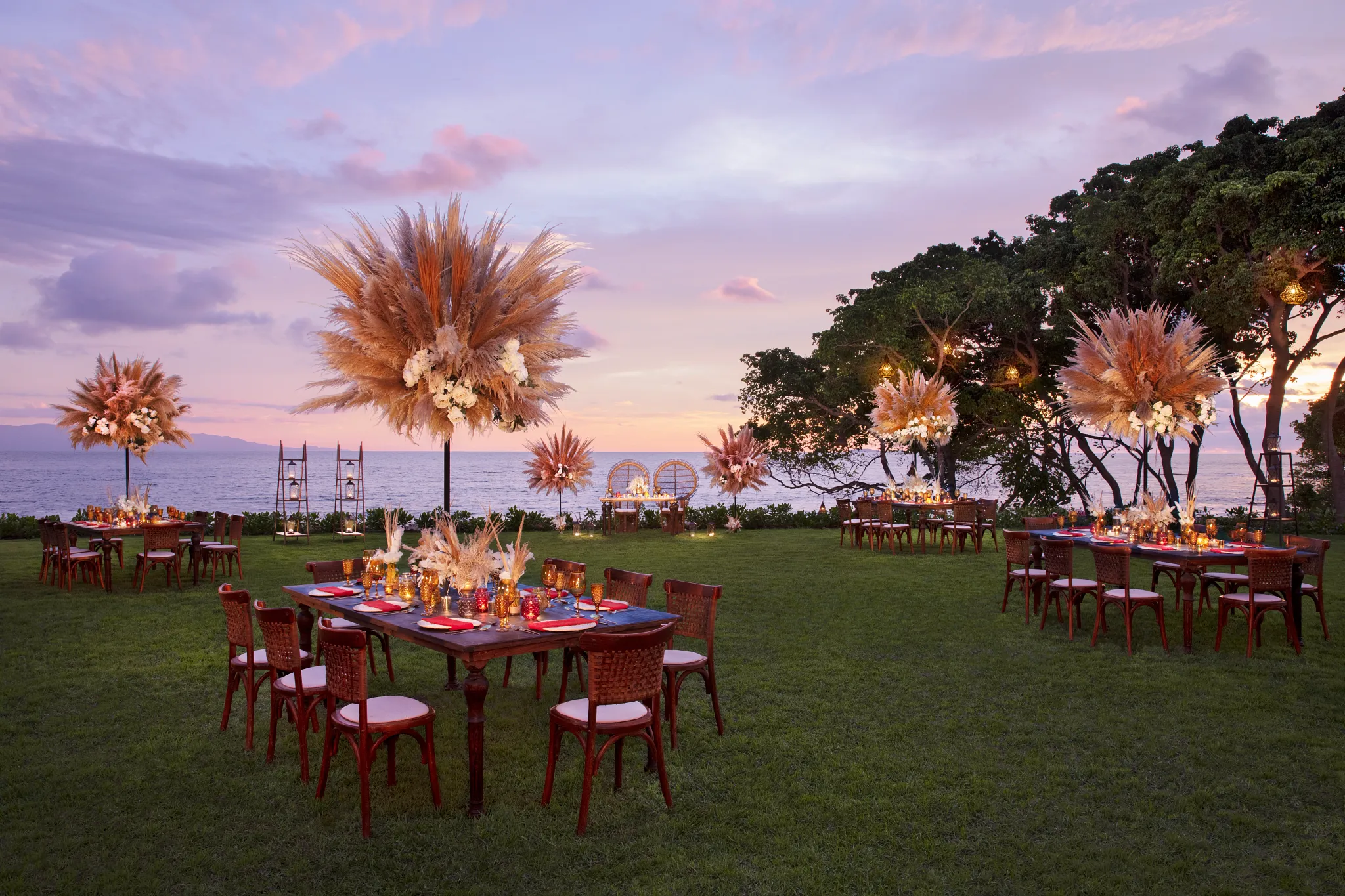 drbmiwedgarden3acb be all inclusive Beachfront outdoor wedding setup at sunset with tall pampas grass arrangements, round tables, chairs, and candles.