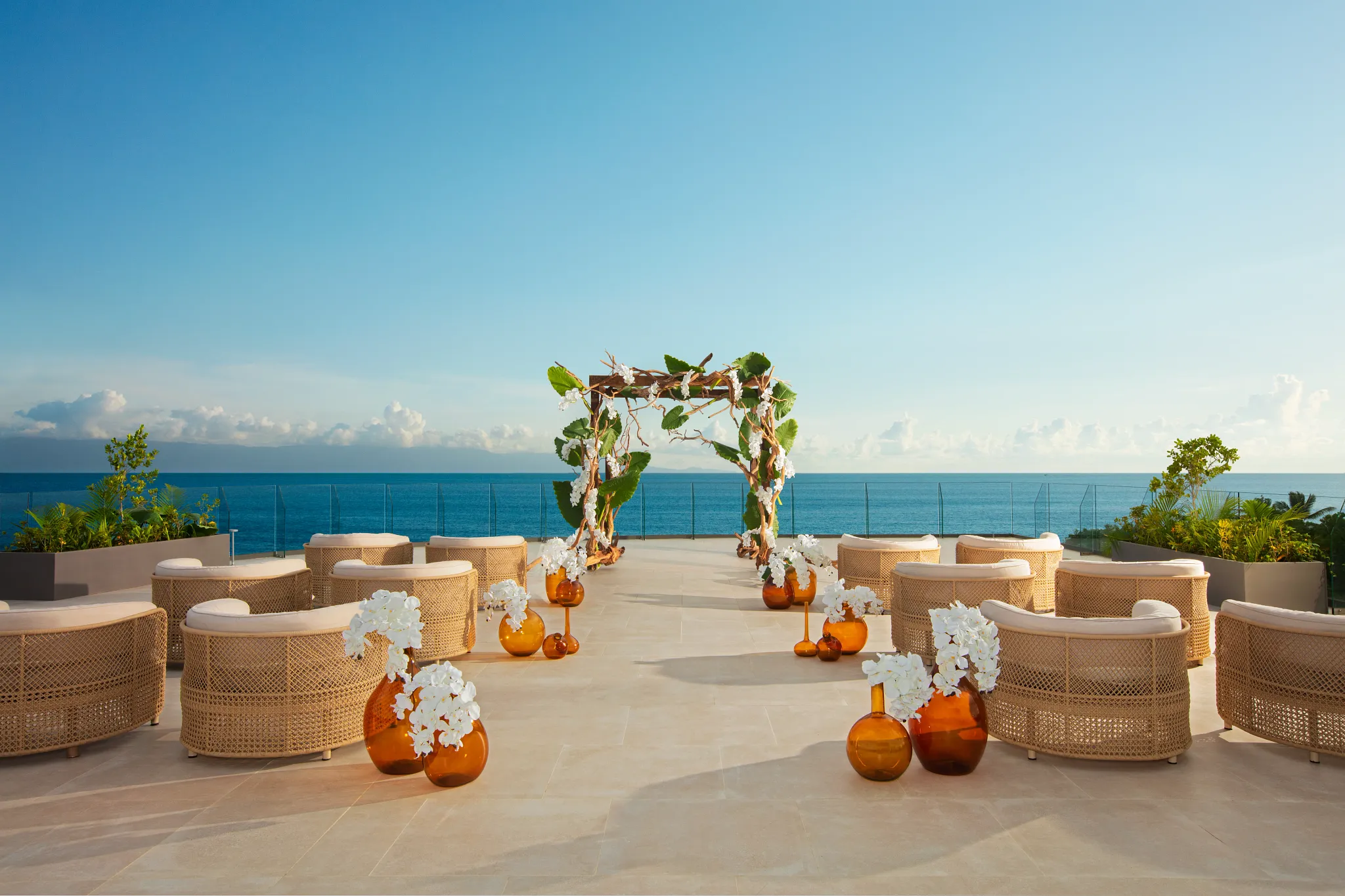 drbmiwedprester1acb be all inclusive Outdoor oceanfront wedding ceremony setup with wicker chairs, floral arch, and orange glass vases, blue sea in the background.