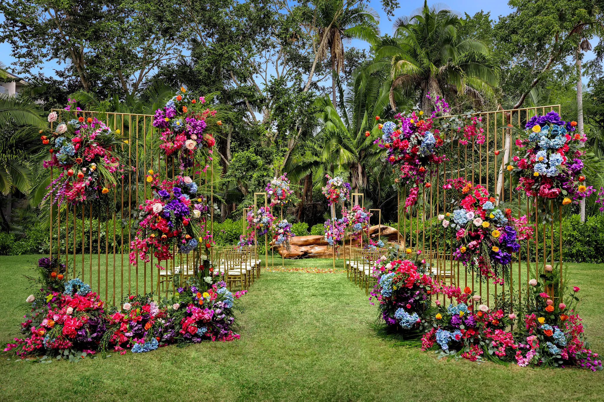 drbmiwedreceptiongarden01afc be all inclusive Outdoor wedding aisle with tall gold floral arches and vibrant blooms along a green lawn. Aisle seating faces the floral backdrop under a sunny sky.