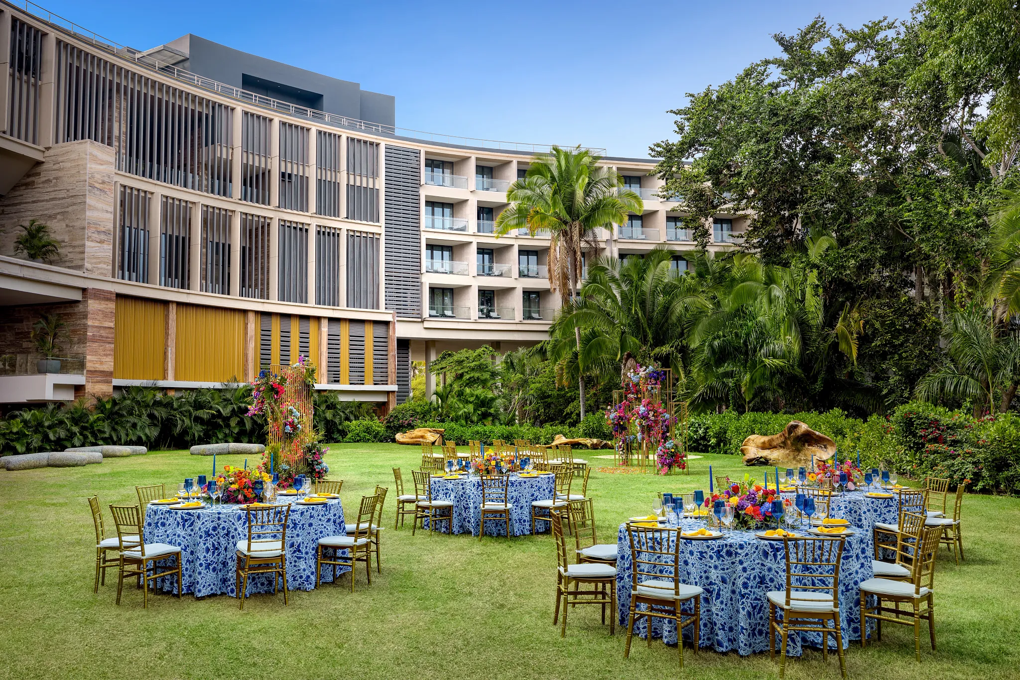 drbmiwedreceptiongarden02afc be all inclusive Outdoor garden banquet with round tables in blue patterned cloths and gold chairs amid tropical landscaping.