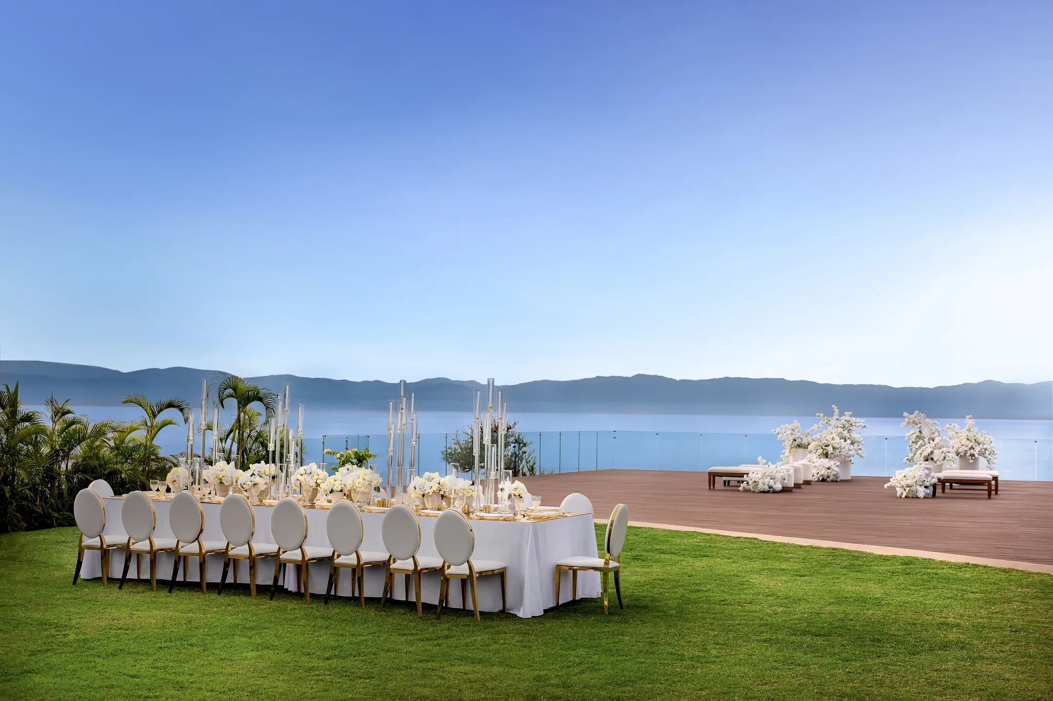 drbmiwedreceptiongardendeck02afc be all inclusive Outdoor wedding reception on a lawn with long white-draped tables, white floral centerpieces, and a glass railing overlooking water and distant mountains.