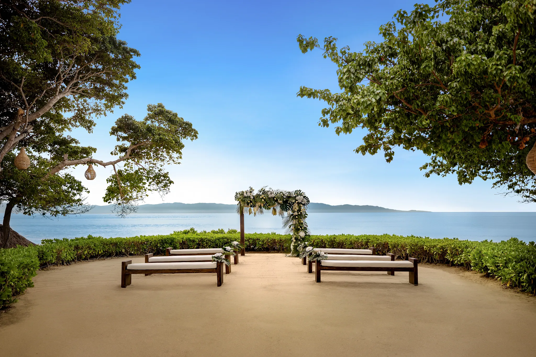 drbmiwedreceptionsandterraceafc be all inclusive Beach wedding ceremony setup with wooden benches and a floral arch overlooking the calm ocean under a blue sky.
