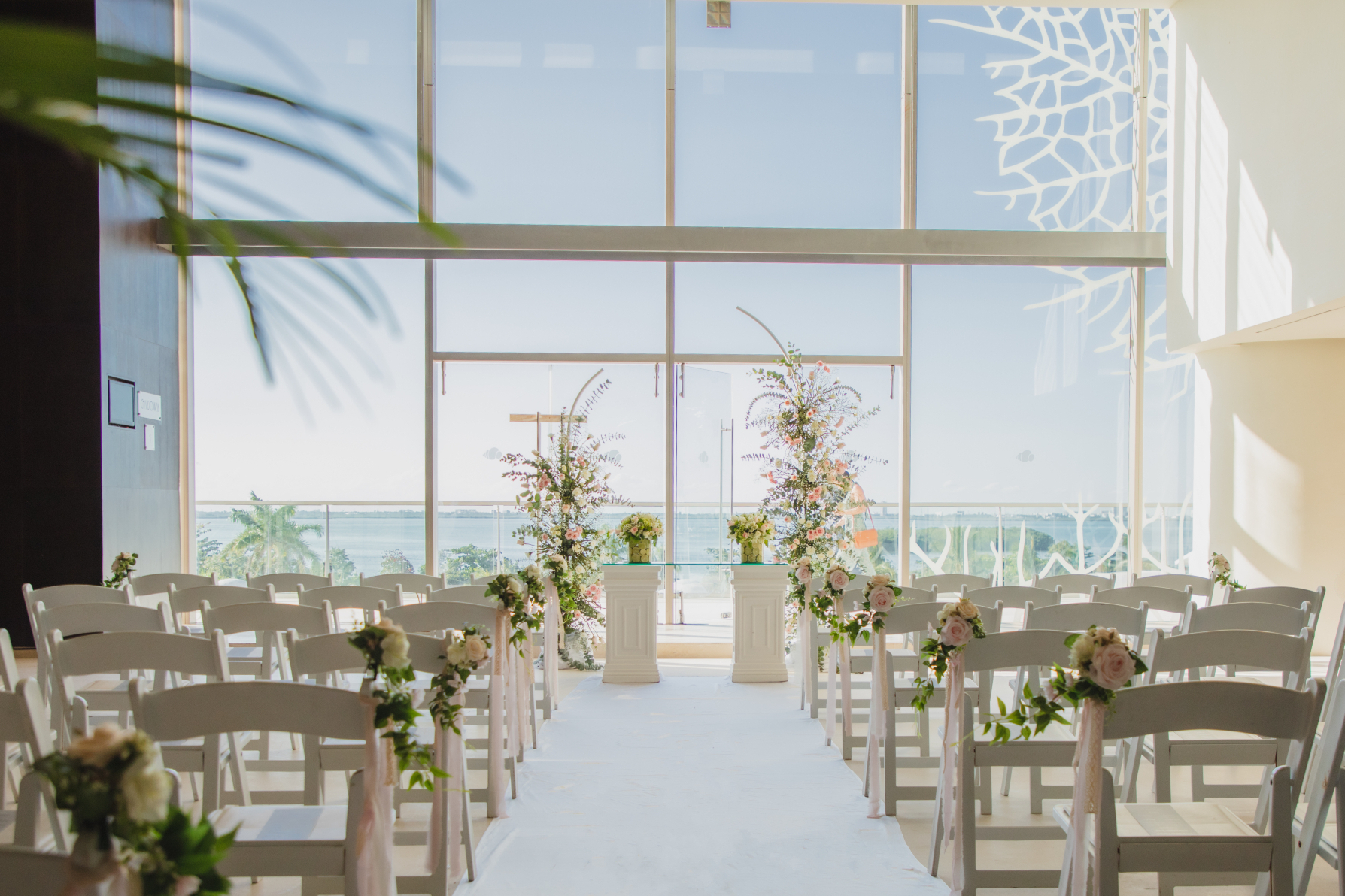 foyer 2 l plan b be all inclusive Bright wedding ceremony space with white chairs lining an aisle, floral arrangements on pedestals, and a glass backdrop overlooking the ocean.