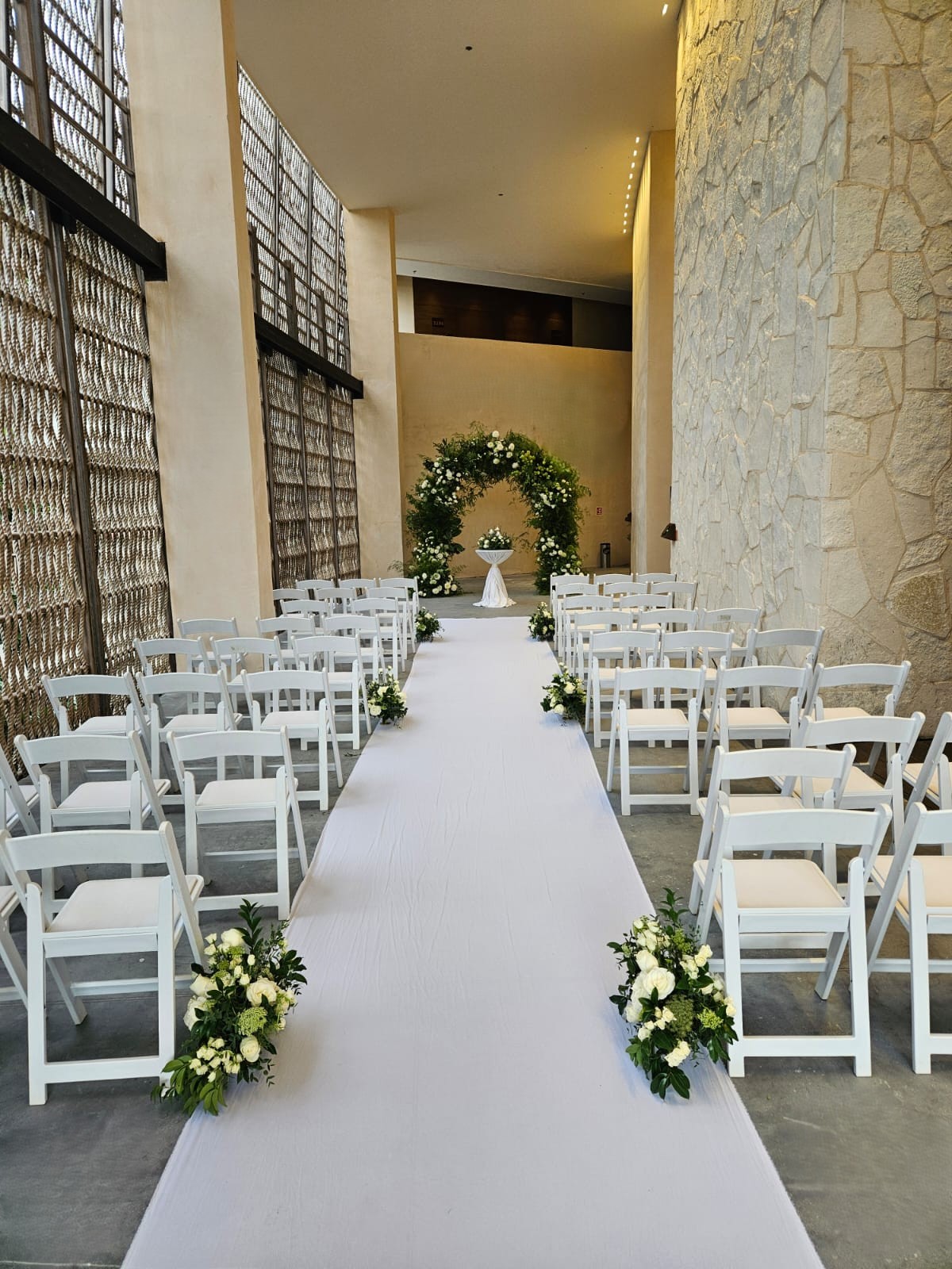 foyer 6 be all inclusive Indoor wedding aisle with white chairs on either side, a white runner, and a floral arch altar at the end in a light stone-walled venue.