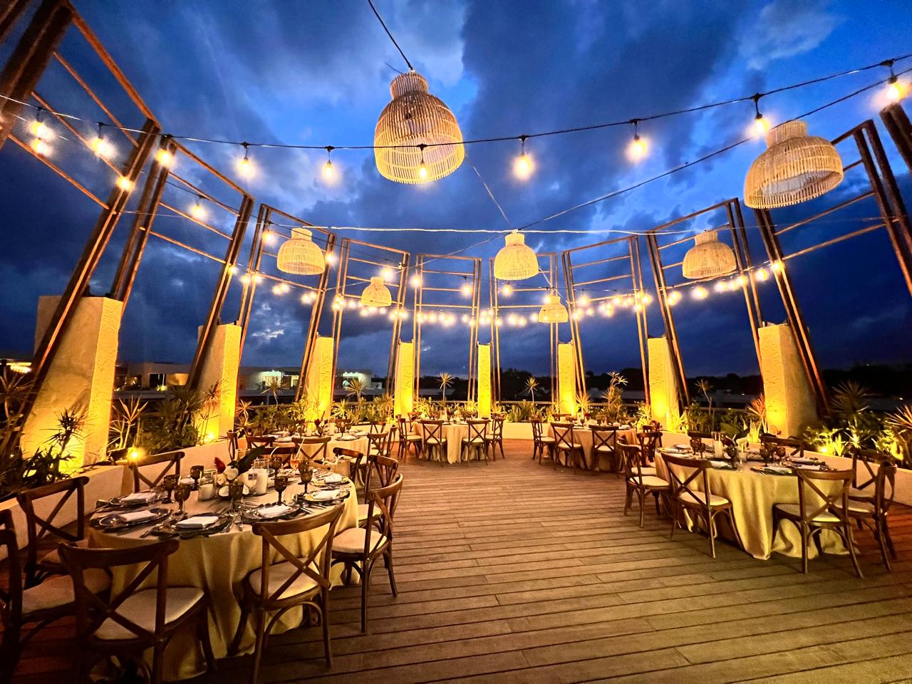 gazebo 1 be all inclusive Outdoor rooftop dining setup at dusk with round tables, warm string lights, wicker lanterns, and wooden chairs.
