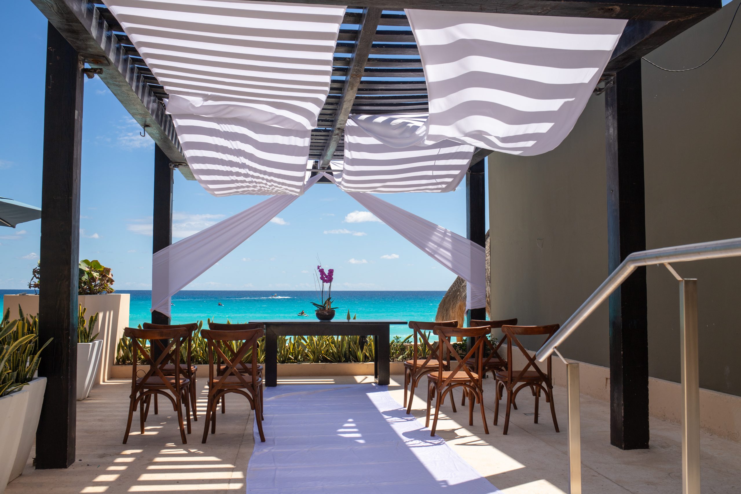gazebo 2 be all inclusive Outdoor seaside dining area with white fabric-draped canopy, wooden chairs, and a blue ocean in the background.