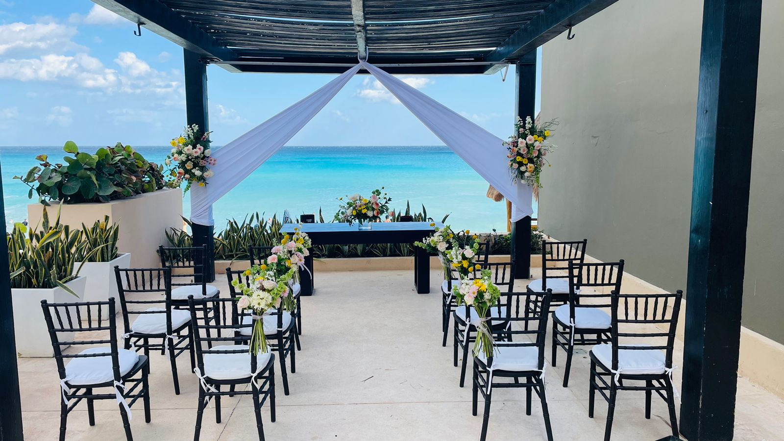 gazebo 6 be all inclusive Beach wedding setup under a dark pergola with white drapes forming an arch, floral bouquets, and rows of black chairs with white cushions facing a table by the turquoise sea.