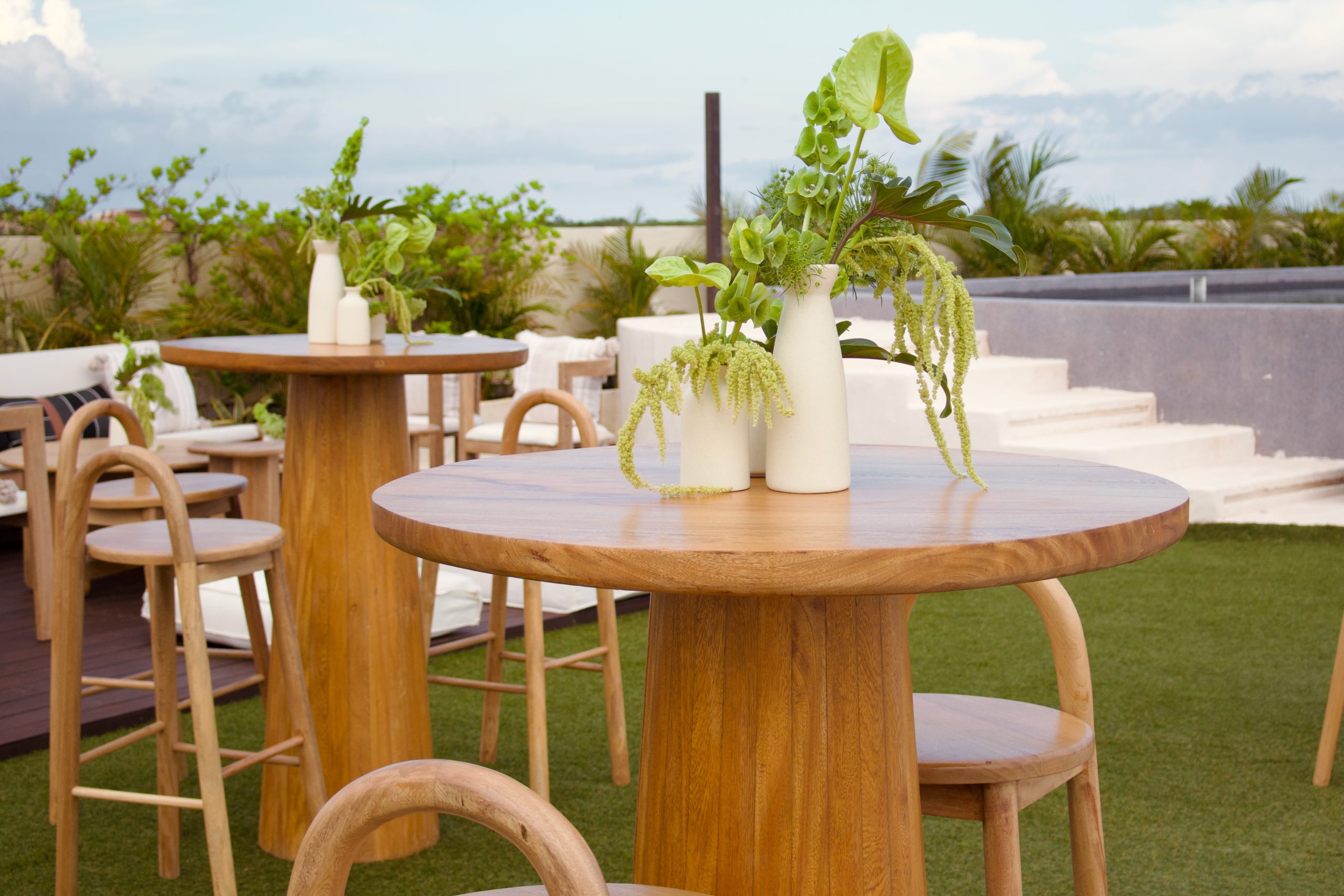 img5456 be all inclusive Outdoor rooftop dining area with round wooden tables, stools, and white ceramic vases filled with greenery