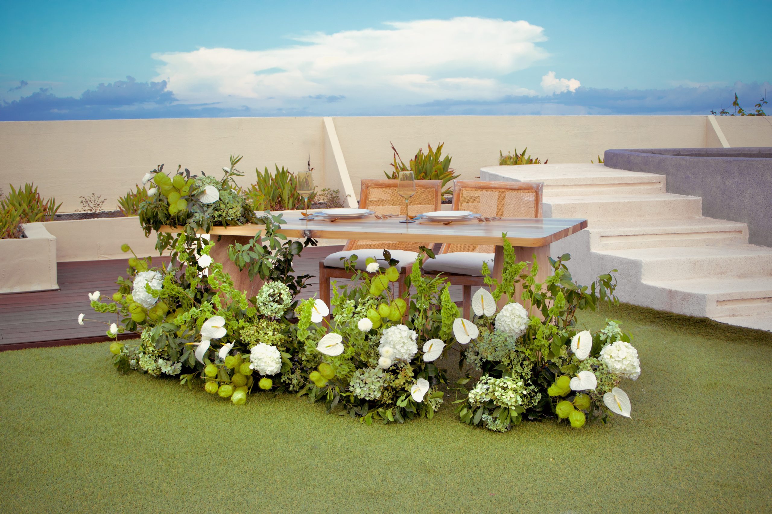 img5461 be all inclusive Outdoor dining table set for two with plates and glasses, surrounded by lush white and green flowers on a patio under a blue sky.