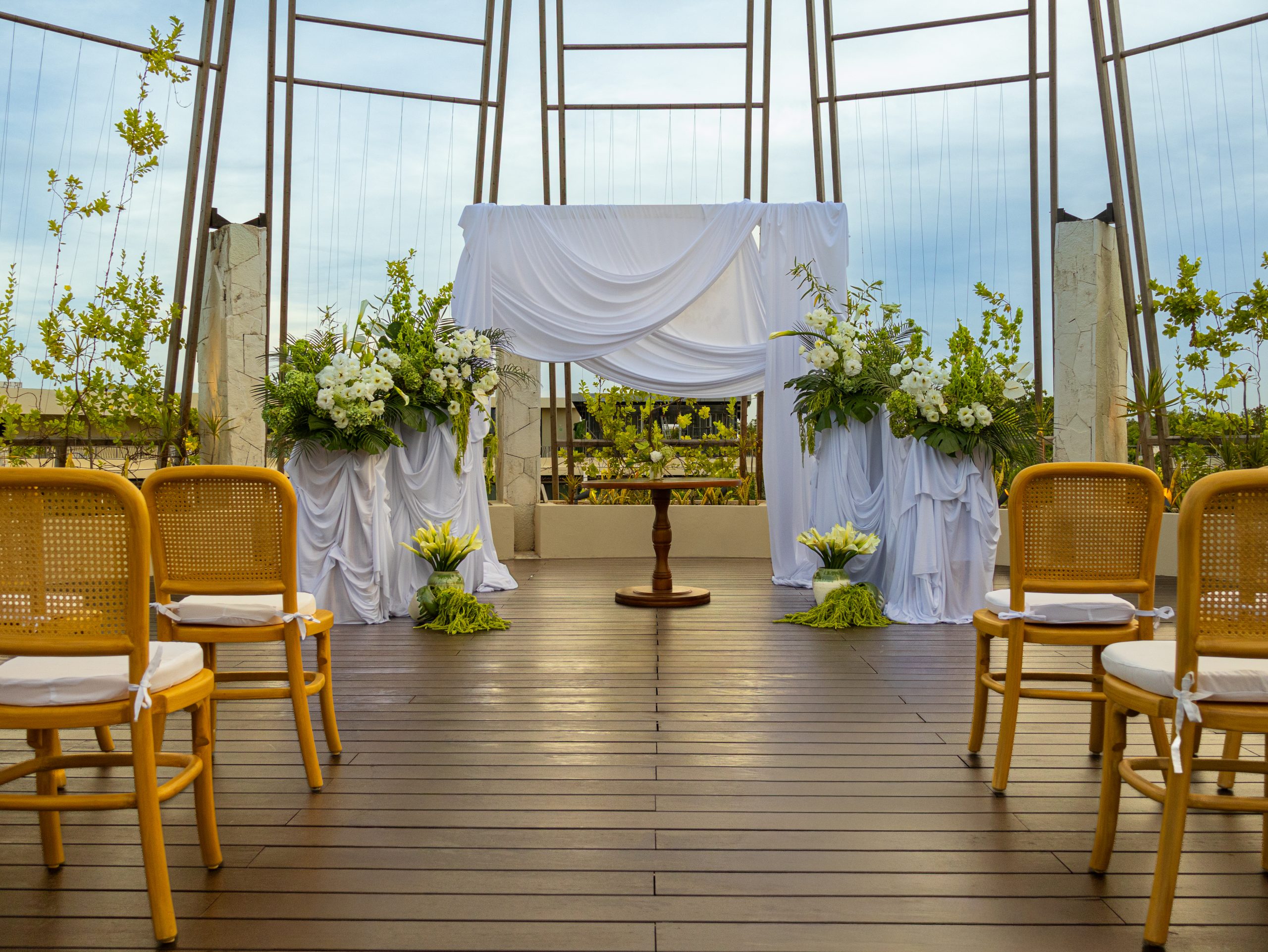 img5510 be all inclusive Outdoor wedding ceremony setup on a wooden deck with a white draped backdrop, floral arrangements, and empty wicker chairs facing the altar.