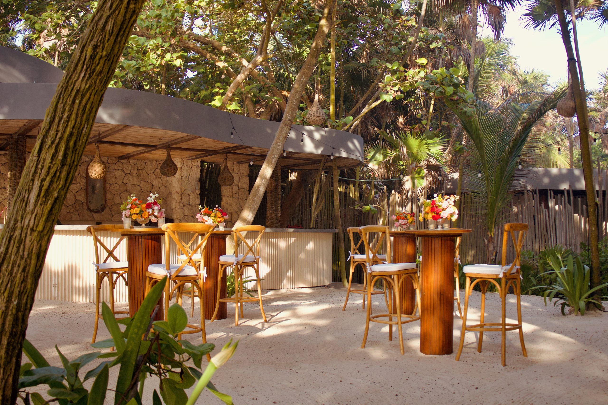 img5544 be all inclusive Outdoor tropical-style seating area with wooden bar tables and chairs, flowering centerpieces, and string lights under lush trees.