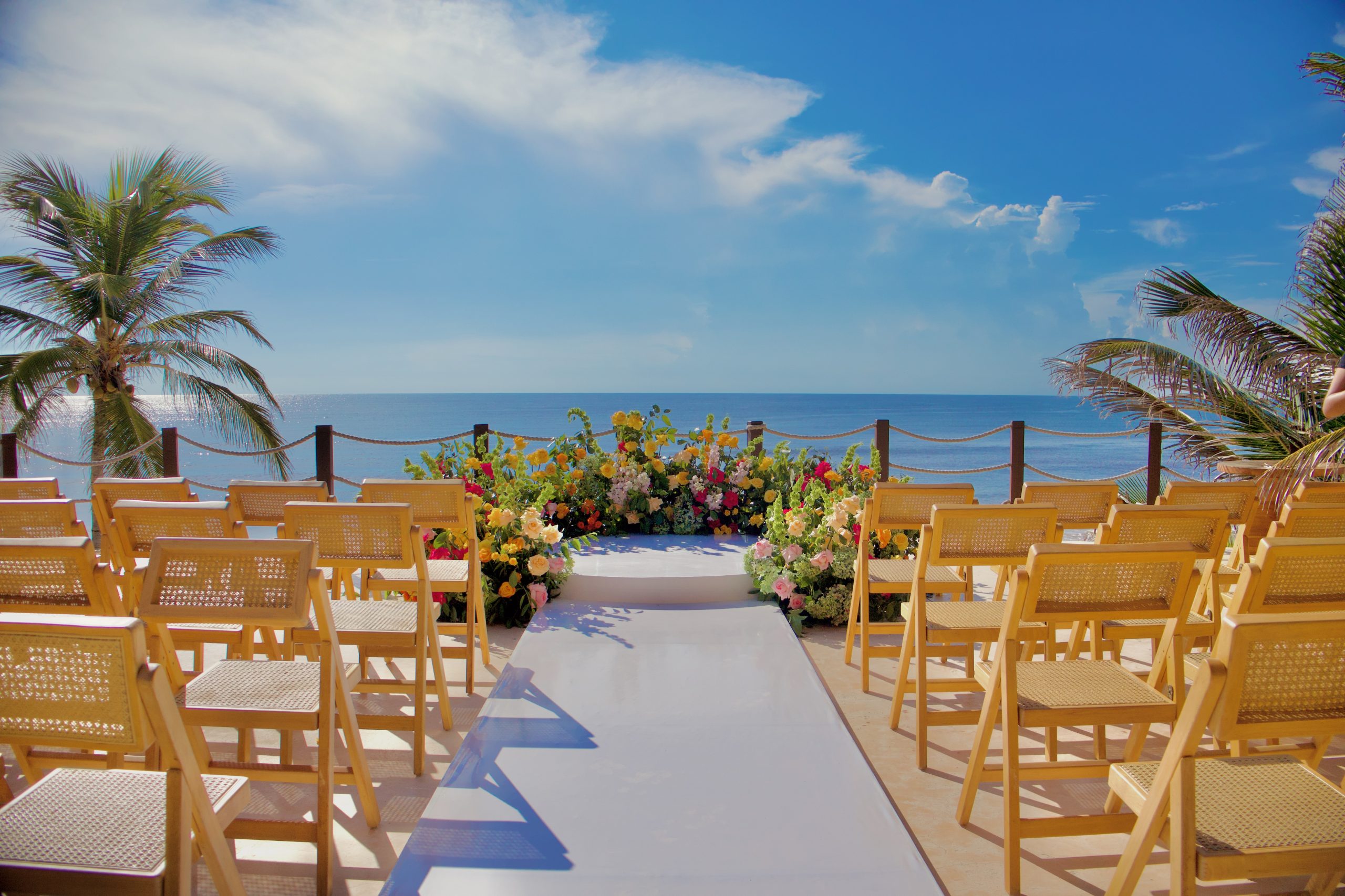 img5623 be all inclusive Outdoor beach wedding setup with yellow chairs facing an ocean view and colorful floral arch at the aisle centerpiece
