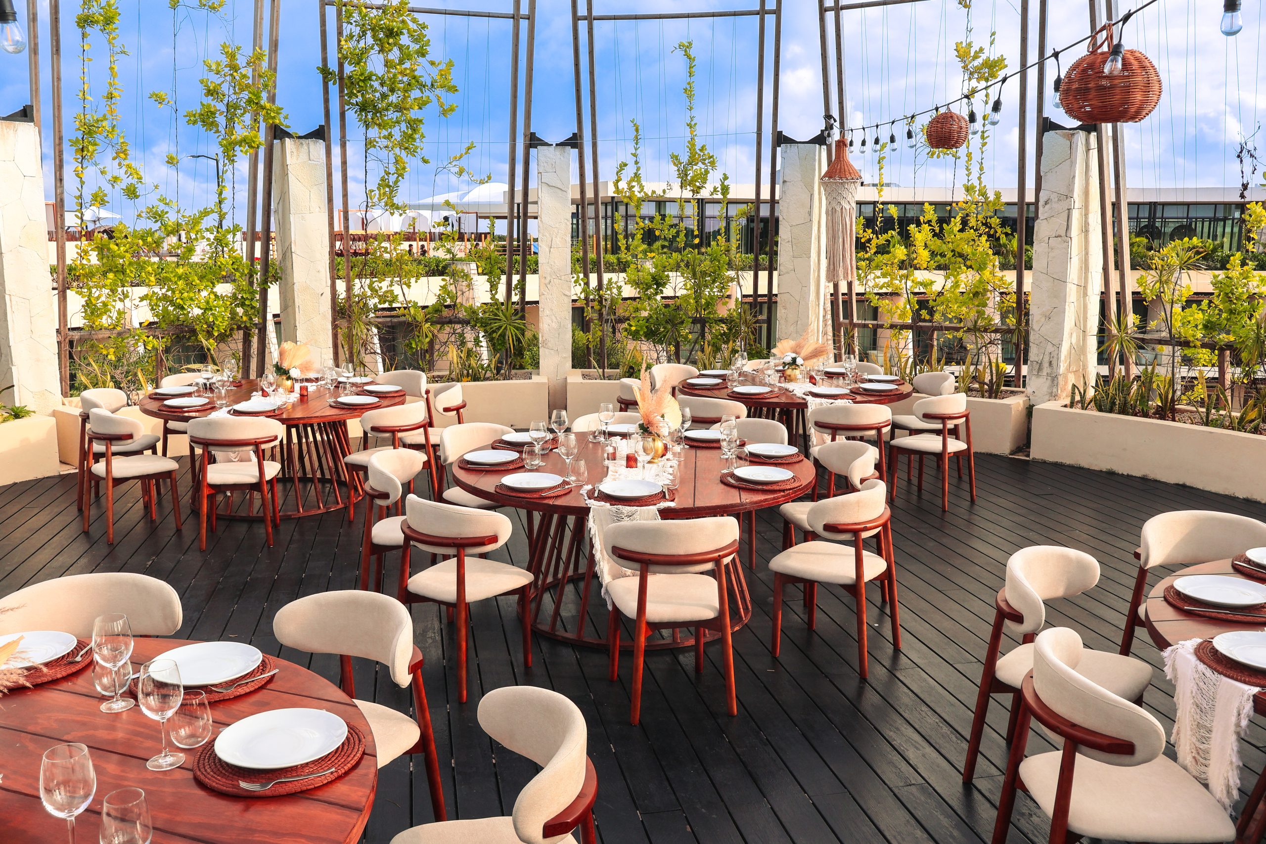 img7222 be all inclusive Outdoor rooftop dining area with round wooden tables set for a banquet, cream chairs, glassware, and greenery against a blue sky.
