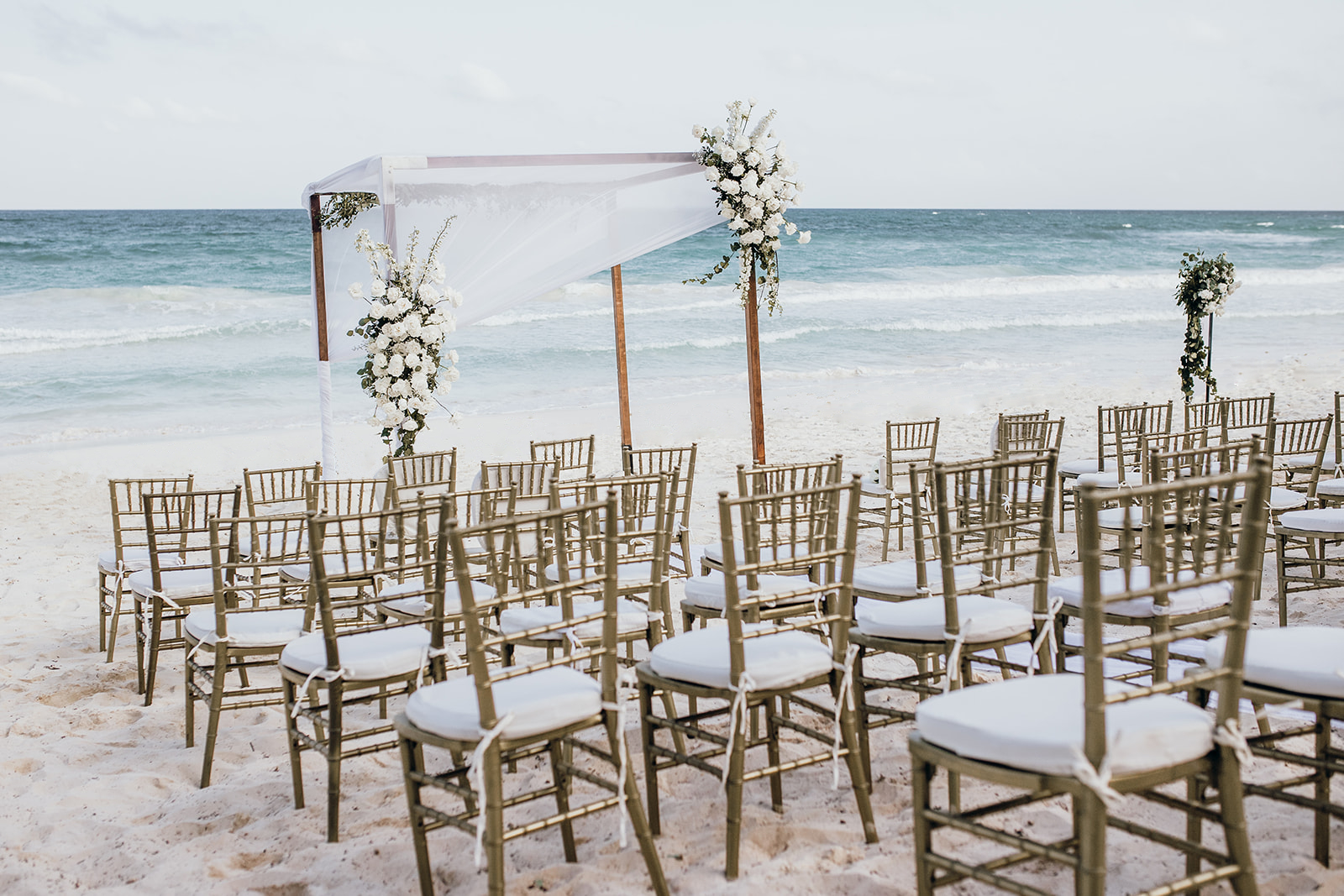secrets tulum be all inclusive Beach wedding ceremony setup with a white-draped arch and floral arrangements overlooking the ocean, rows of gold chairs with white cushions on the sand