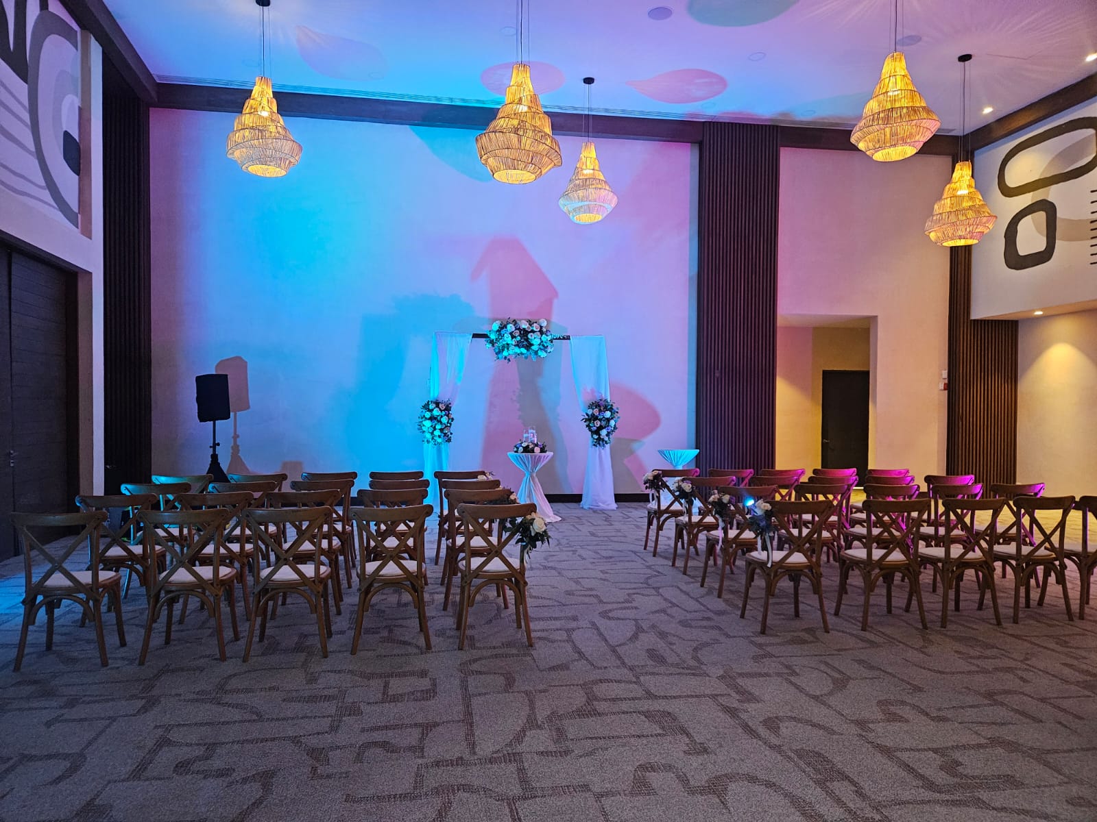 jaguar b 21 be all inclusive Indoor event hall set up for a wedding or ceremony with a floral arch, white drapes, and rows of wooden chairs bathed in blue and purple lighting