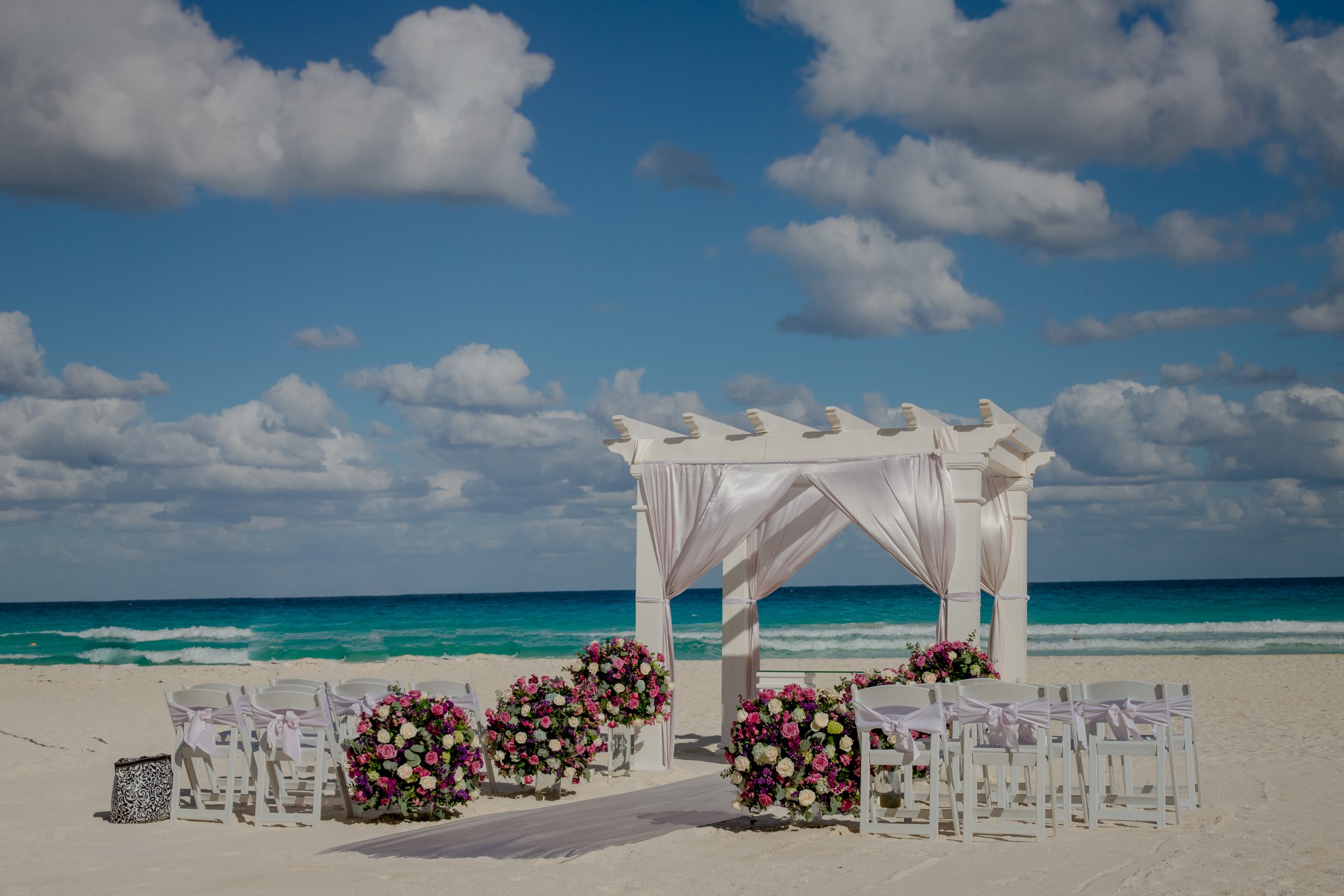 latosha dan2 be all inclusive White wedding arch with flowing white fabric and colorful floral centerpieces on a sandy beach, with the blue ocean in the background.