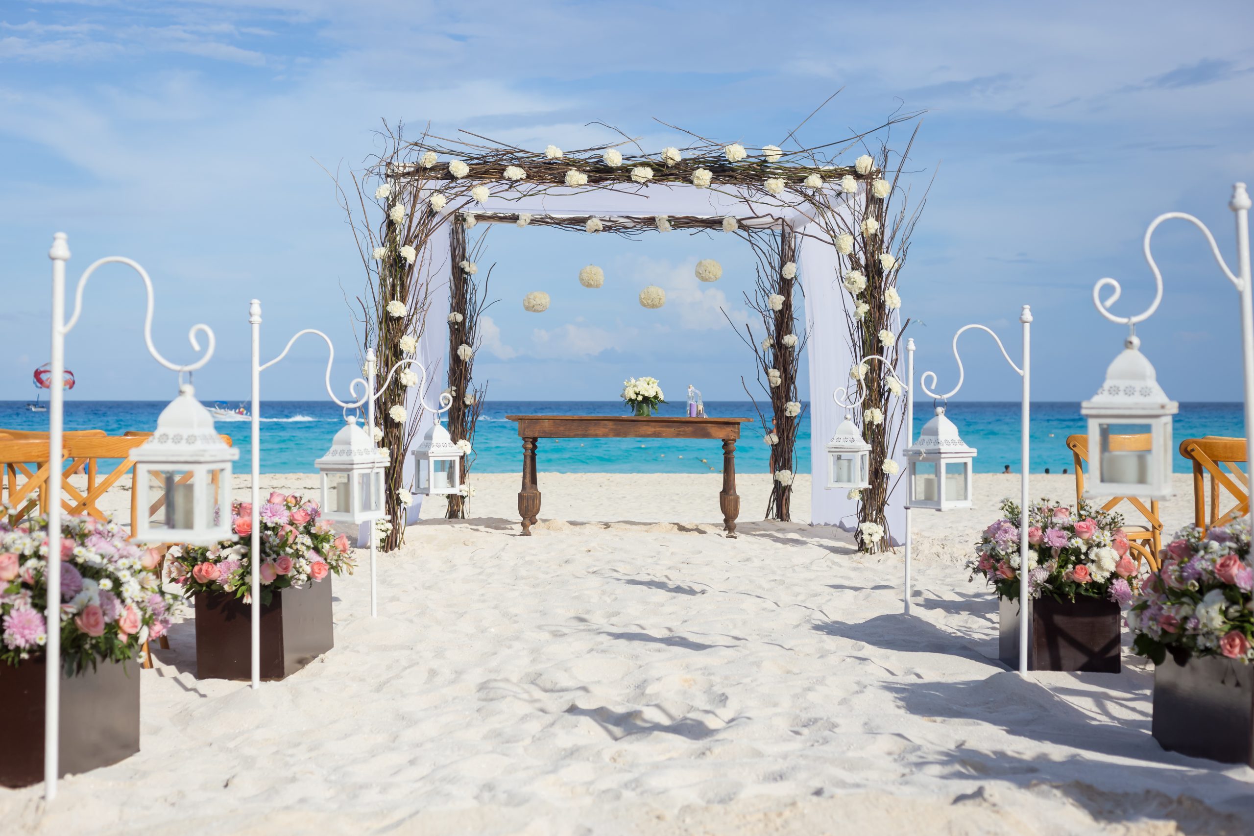 playa 1 be all inclusive Beach wedding altar on the sand with a flower-adorned wooden arch and hanging lanterns, blue sea and sky in the background.