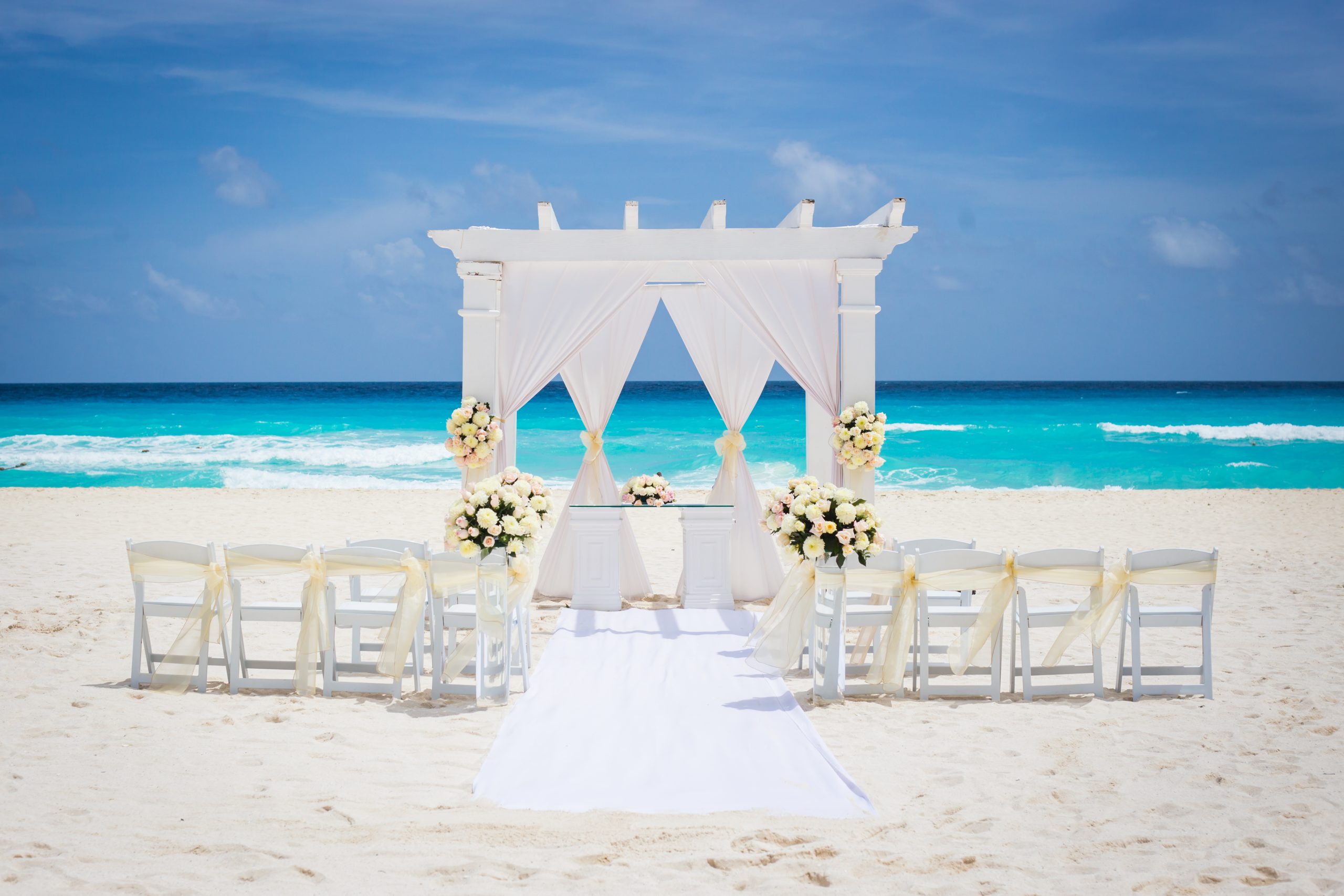 playa 2 be all inclusive White wedding arch on a sandy beach, draped with white fabric and floral arrangements, facing the turquoise sea.