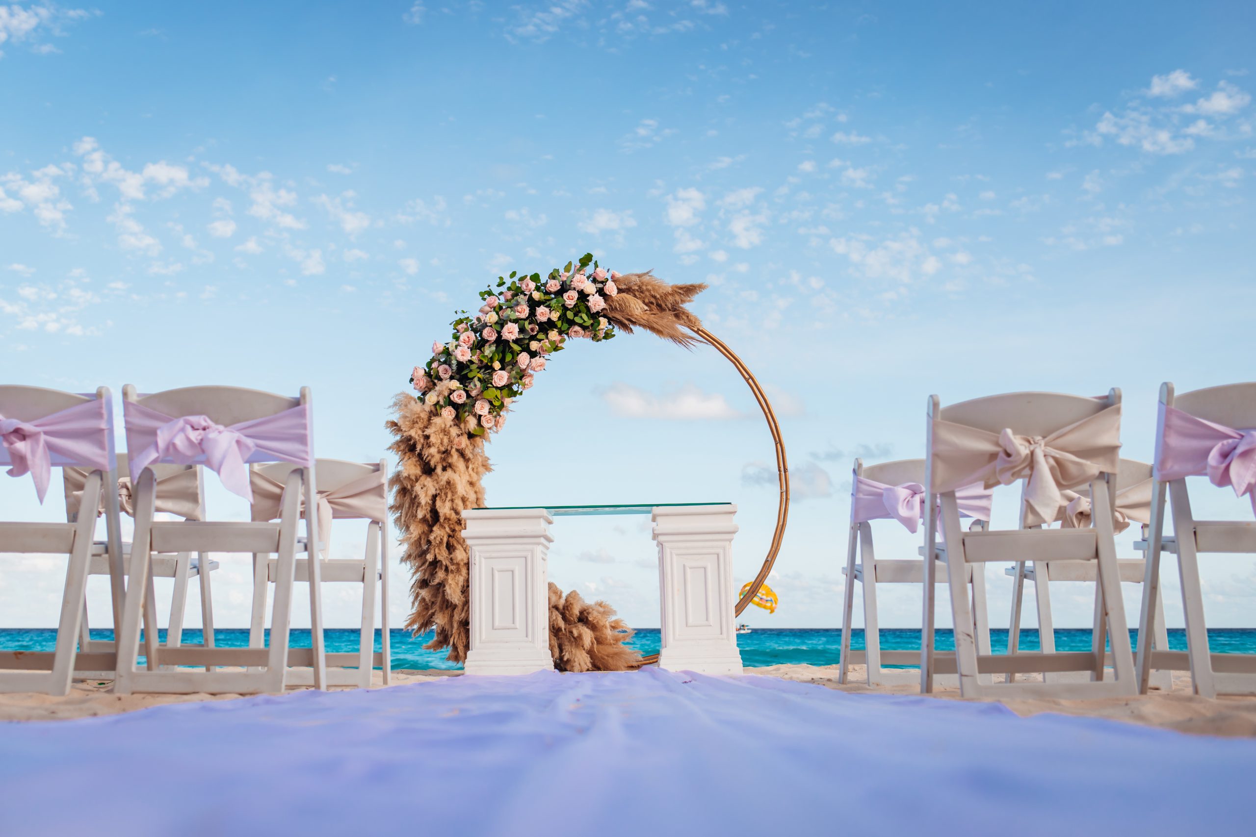 playa 8 be all inclusive Beach wedding setup with a floral arch, white chairs with pastel ribbons, and a blue ocean backdrop.