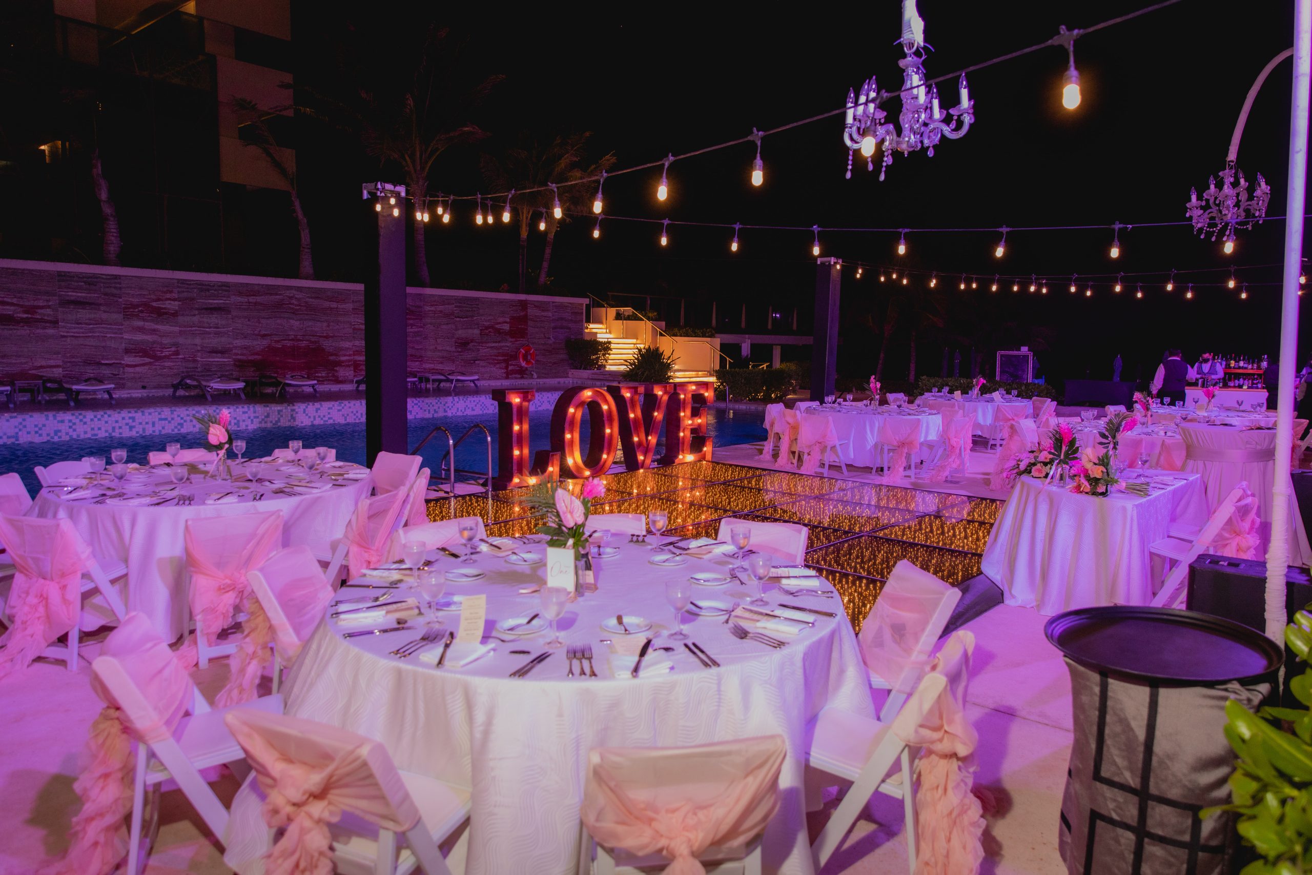 kristyna prashanth be all inclusive Outdoor nighttime wedding reception by a pool with string lights, pink/purple lighting, and illuminated 'LOVE' sign near tables set for guests.