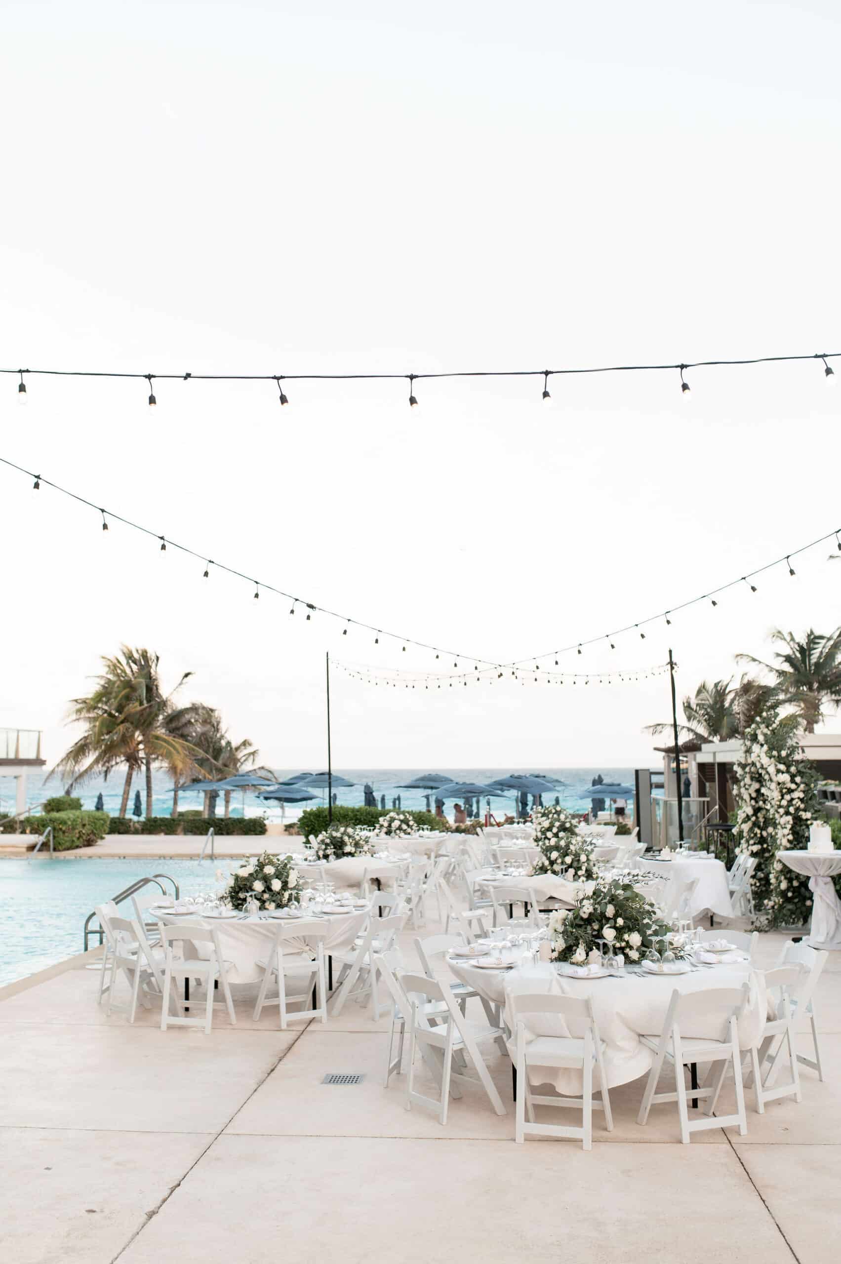 pool side terrace 7 be all inclusive Outdoor beachside event with white-draped tables and chairs, floral centerpieces, and string lights over a seaside setting