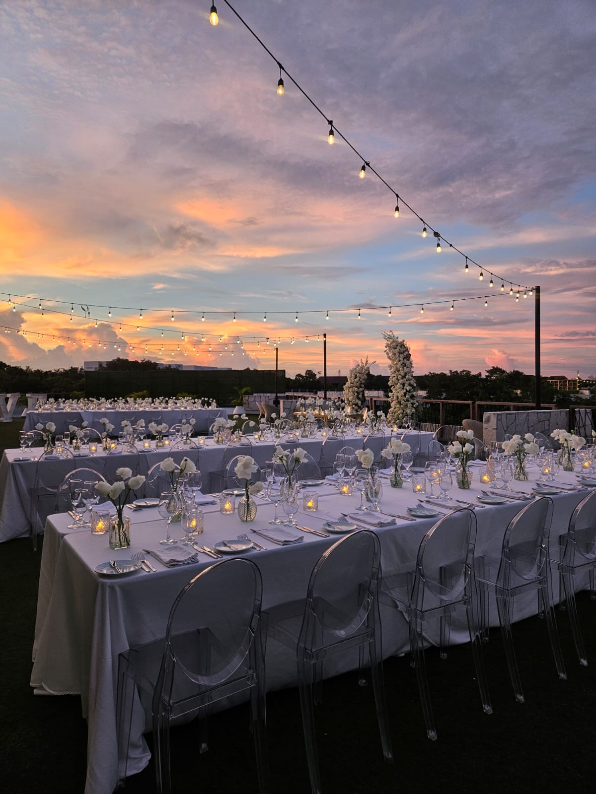 rooftop garden 3 be all inclusive Outdoor evening wedding reception with long white-draped tables, floral centerpieces, candles, and string lights against a sunset sky.