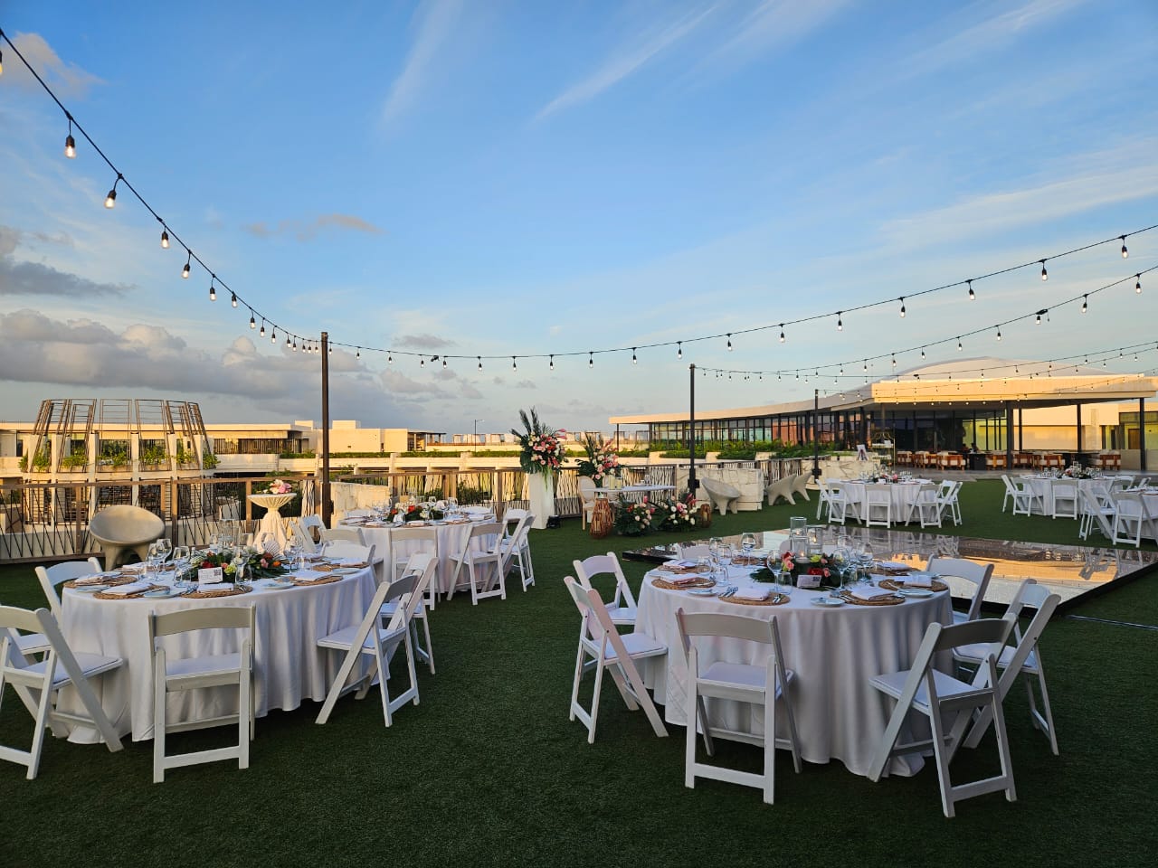 rooftop garden 5 be all inclusive Outdoor wedding reception on a grassy terrace with round tables, white chairs, and string lights against a blue sky.