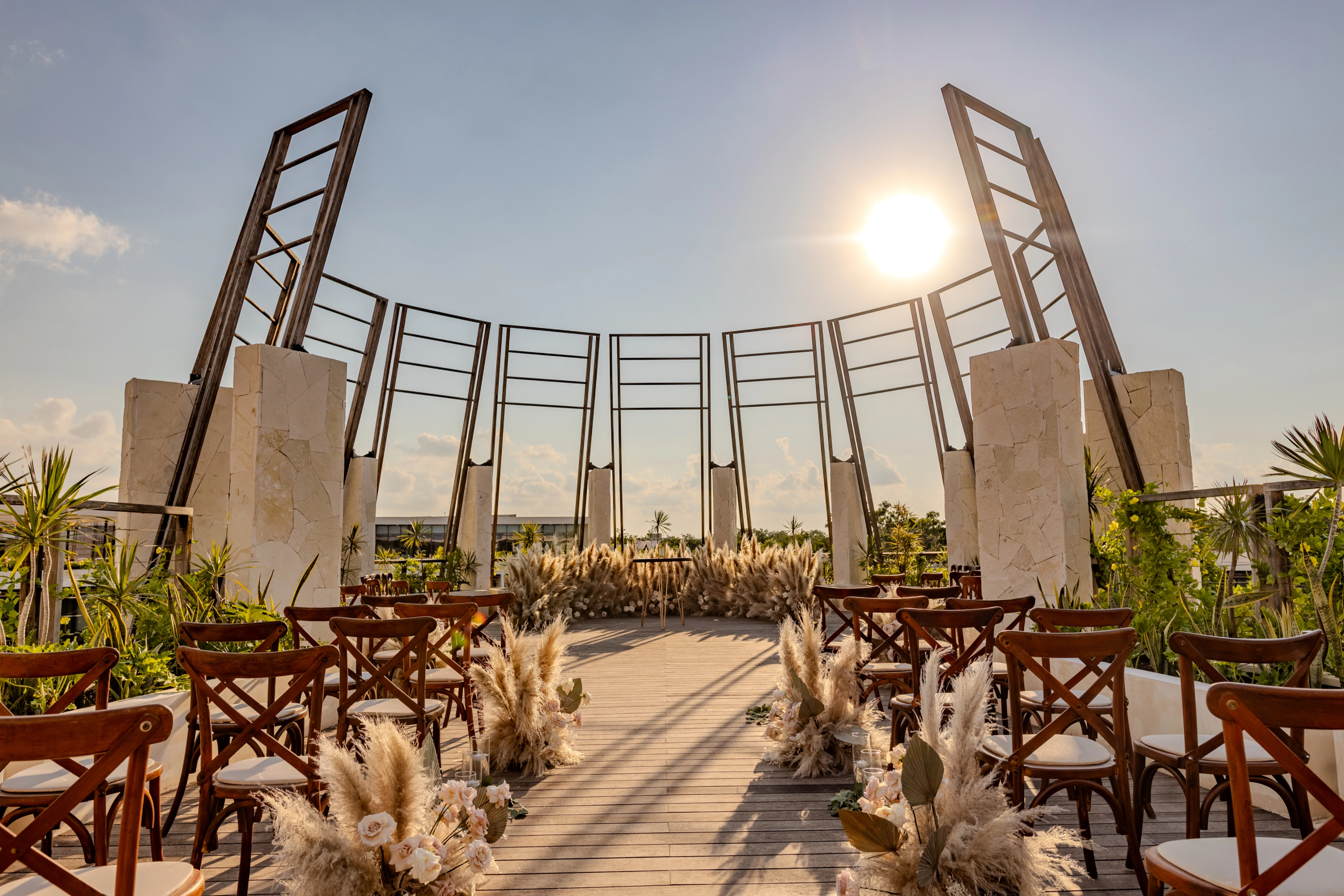 sectuwedskygazebo01afc be all inclusive Outdoor wedding ceremony setup with wooden chairs arranged around a stone archway and tall wooden frames under a sunny sky, tropical plants in the foreground.