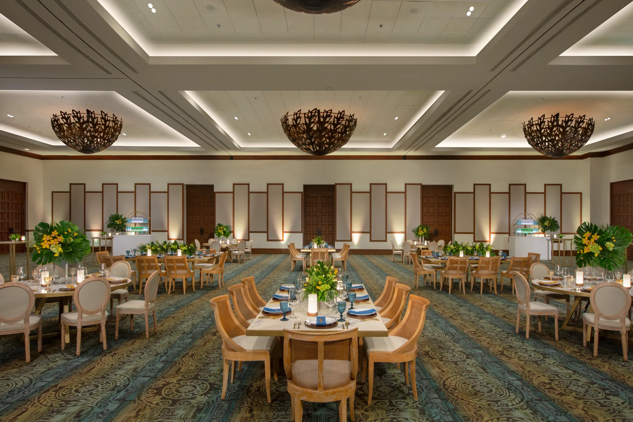semrcgrpballroom3acb be all inclusive Elegant banquet hall set for a formal event with sculpted wooden chandeliers, arranged tables, blue napkins, glassware, and yellow floral centerpieces on a patterned carpetled room.