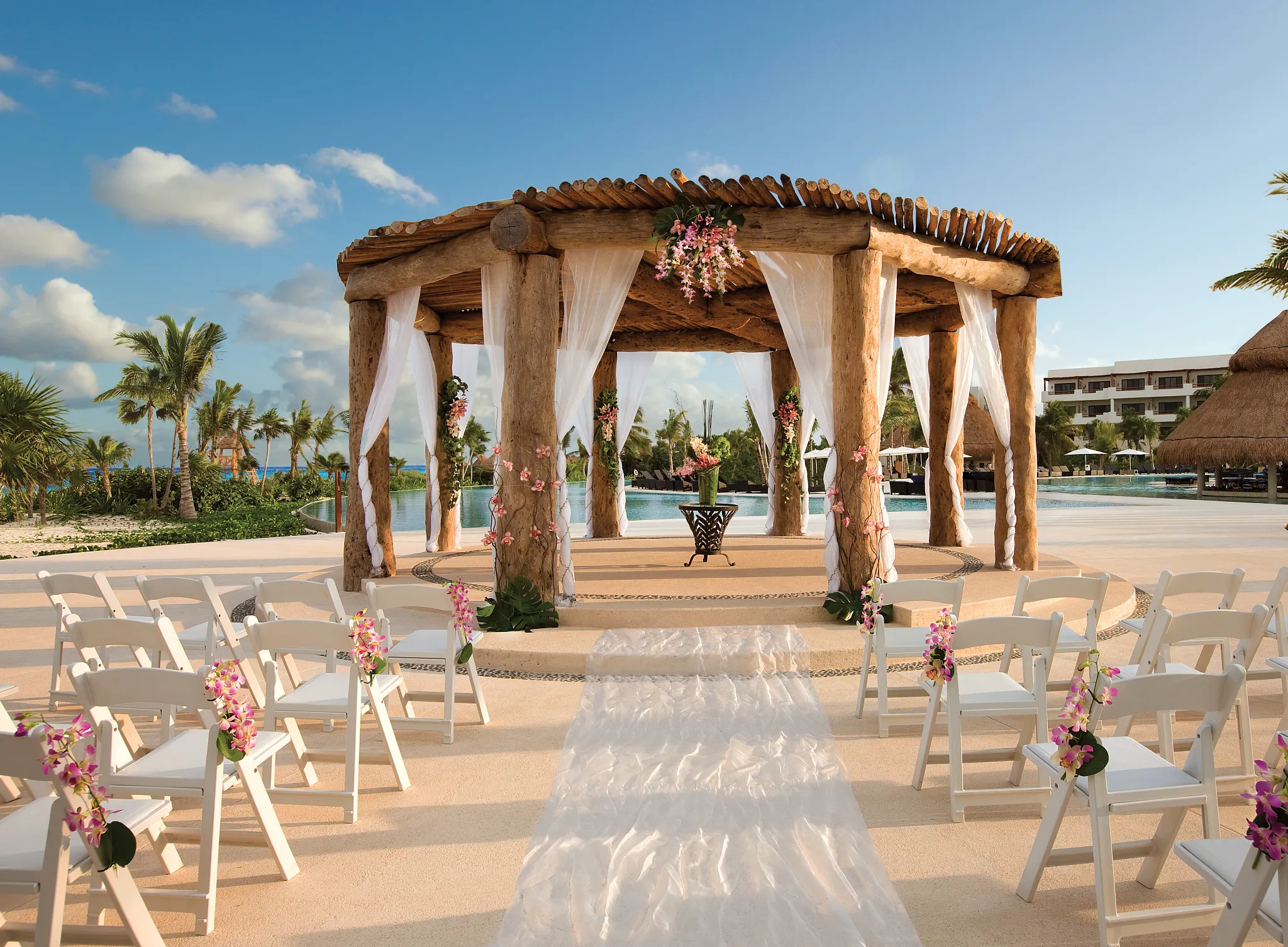 semrcwedwedding gazebo2cb be all inclusive Beach wedding setup with a wooden arch, white drapes, and pink floral decorations along an aisle.