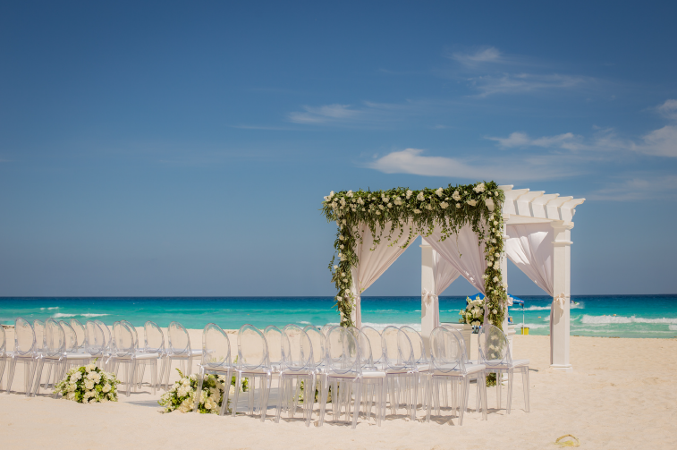 screenshot 20260609 115637 be all inclusive White seaside wedding arch with greenery and sheer drapes on a sandy beach, clear chairs arranged for ceremony with ocean backdrop