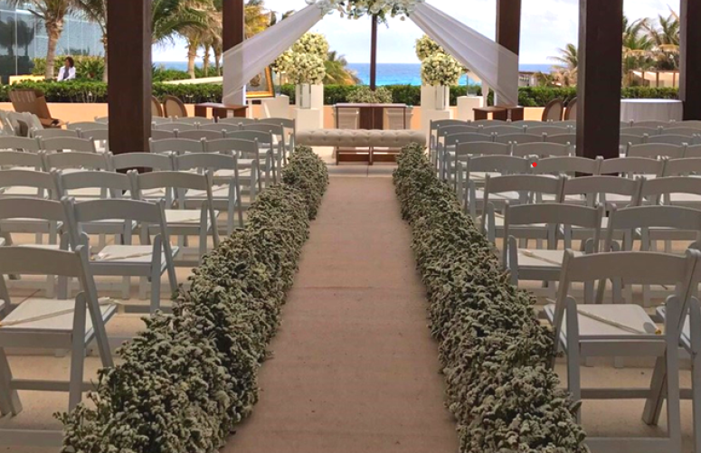 screenshot 20260609 115738 be all inclusive Outdoor wedding ceremony setup: an aisle flanked by white chairs and flower-adorned hedges leading to a draped arch with the ocean in view.