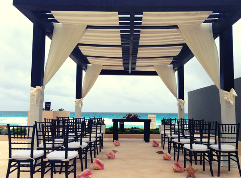 screenshot 20260609 120037 be all inclusive Beach wedding setup under a dark pergola with flowing white drapes, rows of black chairs, and pink seashells along the aisle. The ocean is in the background.