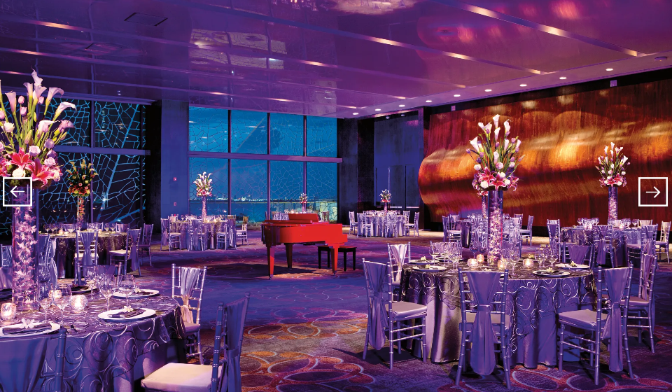 screenshot 20260609 120824 be all inclusive Event ballroom with round tables, purple lighting, tall floral centerpieces, and a red grand piano in the background