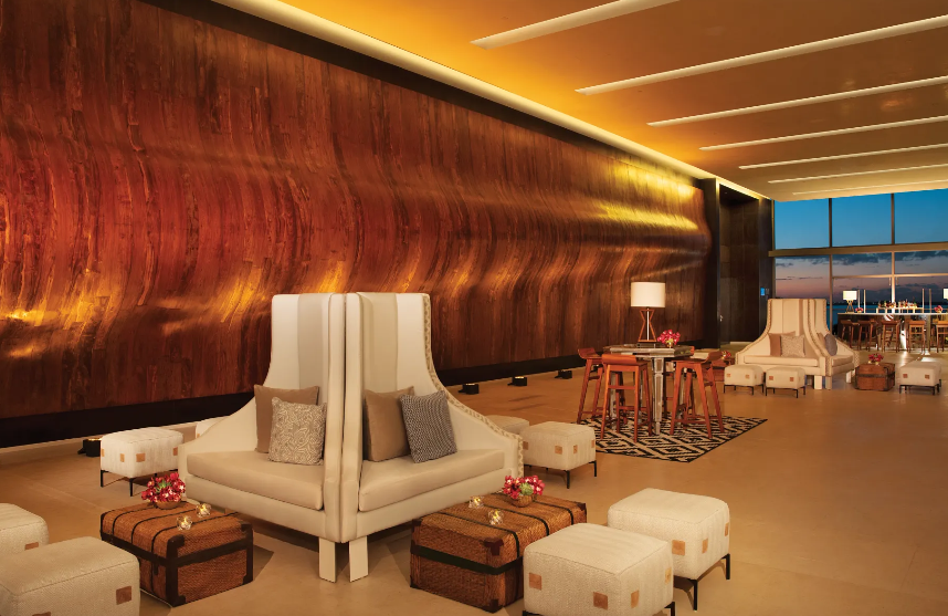 screenshot 20260609 120903 be all inclusive Luxurious hotel lounge with tall beige armchairs, ottomans, and warm wood wall under soft lighting, sunset visible through windows.