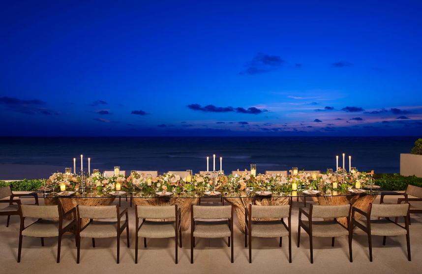 screenshot 20260609 120926 be all inclusive Seaside outdoor banquet with a long candlelit table, floral centerpieces, and chairs facing the ocean at dusk.