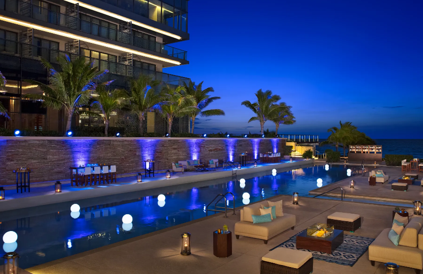 screenshot 20260609 121006 be all inclusive Nighttime poolside at a resort with lounge seating, palm trees, and ocean view in the distance.