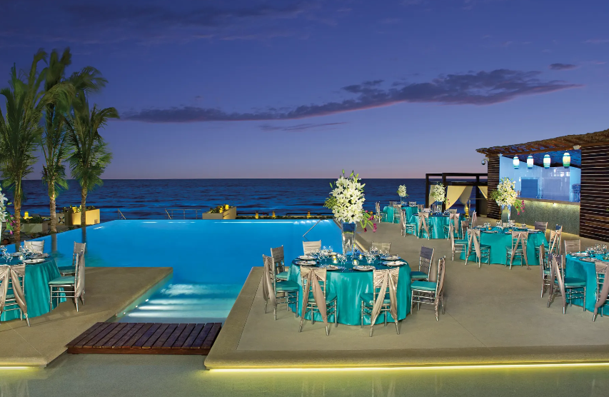 screenshot 20260609 121101 be all inclusive Poolside banquet setup with turquoise tablecloths, white floral centerpieces, and elegant dining chairs against an ocean horizon at dusk.