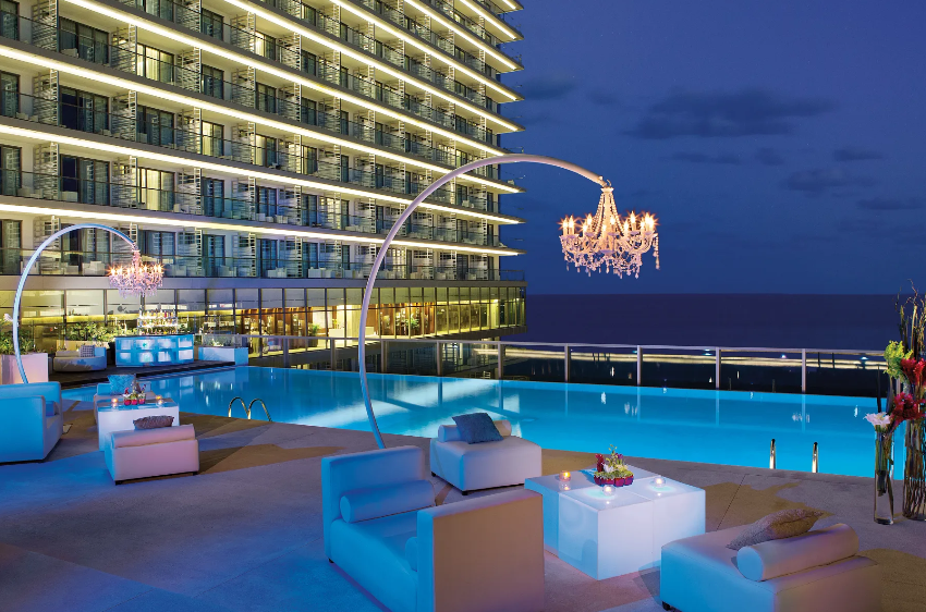 screenshot 20260609 121126 be all inclusive Rooftop pool area at night with curved chandeliers, white lounge seating, and an ocean view beyond the glass railing.