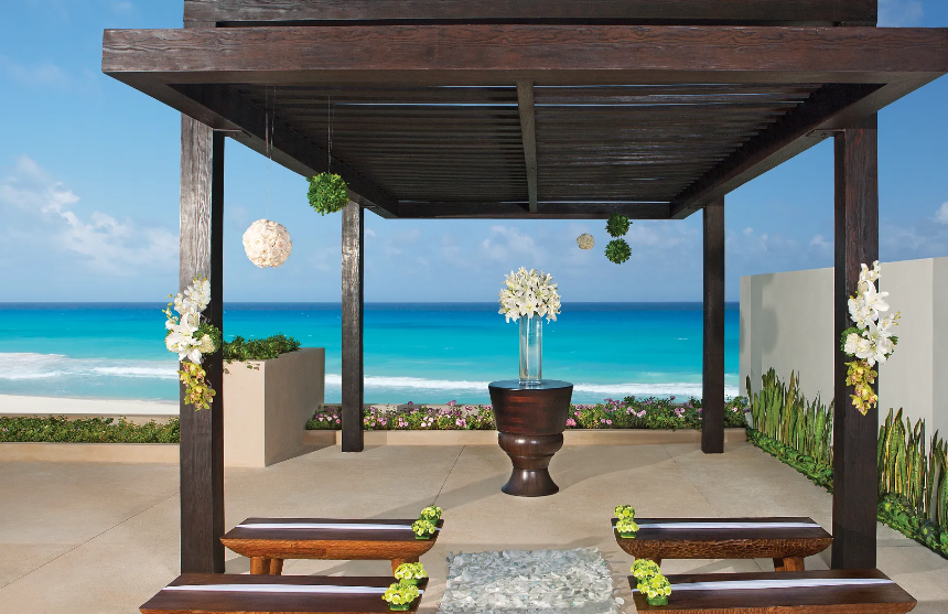 screenshot 20260609 121205 be all inclusive Outdoor beach wedding altar under a dark wooden pergola with white flower arrangements and an ocean backdrop.