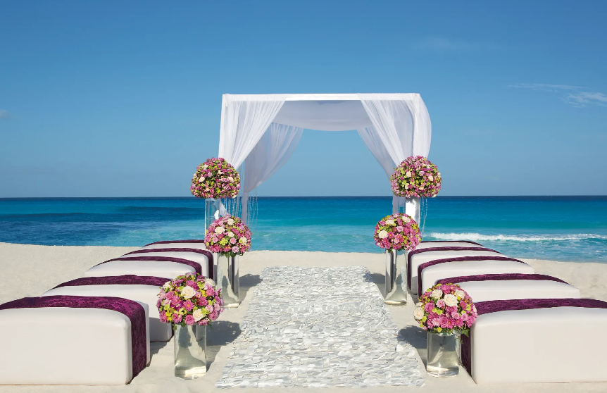 screenshot 20260609 121236 be all inclusive Beach wedding ceremony setup with a white draped arch and pink flower bouquets along the aisle, facing the ocean.