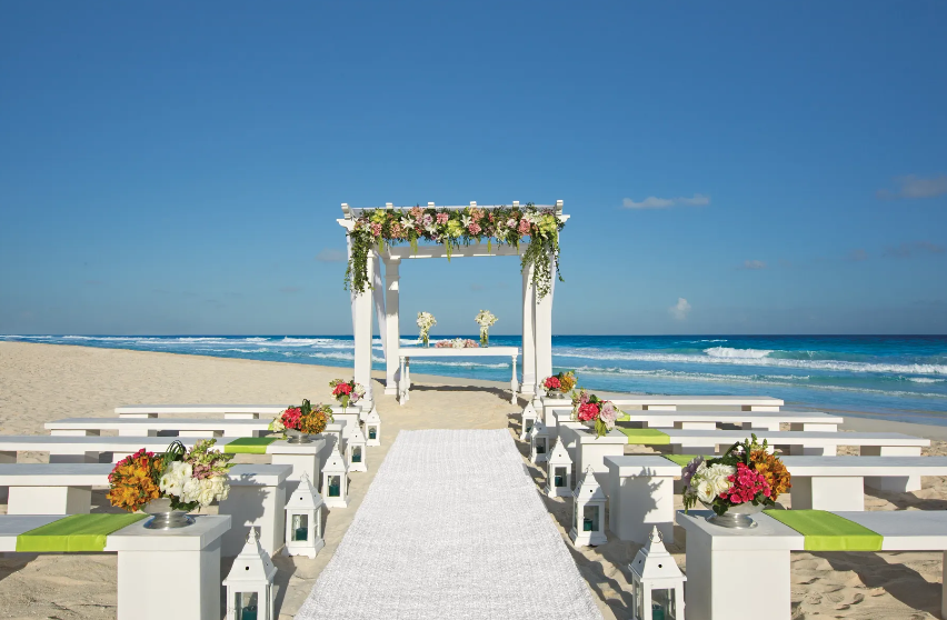 screenshot 20260609 121259 be all inclusive Beach wedding setup with a white floral arch and aisle leading to the ocean beyond.