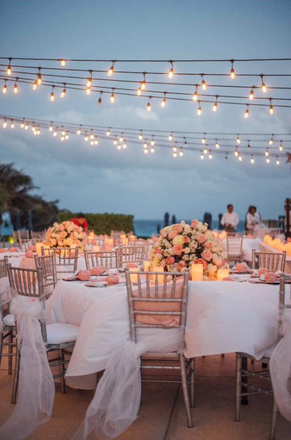 screenshot 20260609 122223 be all inclusive Outdoor evening wedding reception on a beach terrace with string lights, white linens, and pink/peach floral centerpieces with candles.