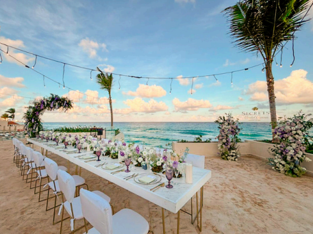 screenshot 20260609 135632 be all inclusive Beach wedding setup: long white-draped table with white chairs, floral centerpieces, string lights, and the ocean in the background on the sandy shore.