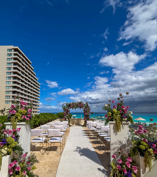 screenshot 20260609 135720 be all inclusive Outdoor seaside wedding ceremony setup on a terrace with white chairs, a flower-decorated arch, and a view of the ocean under a blue sky.