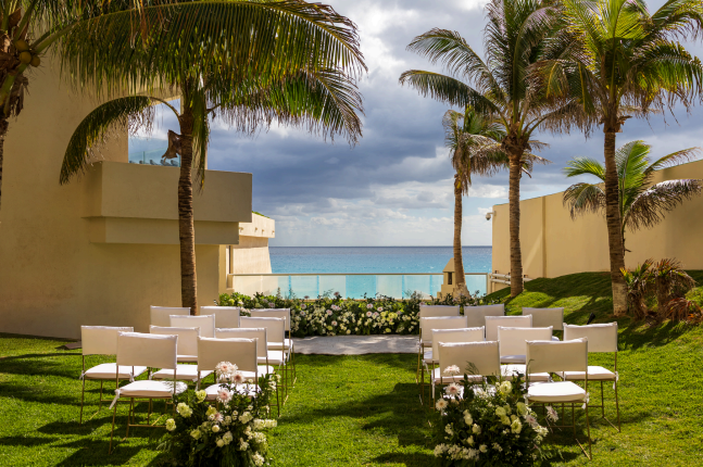 screenshot 20260609 135859 be all inclusive Outdoor seaside ceremony setup with white chairs and floral arrangements, palm trees, and ocean view.