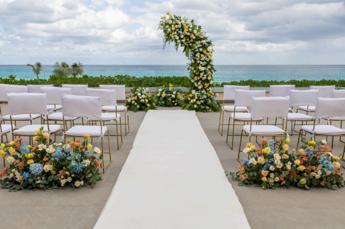 screenshot 20260609 135959 be all inclusive Beach wedding ceremony setup with white chairs along a white aisle runner and a floraled arch overlooking the sea.