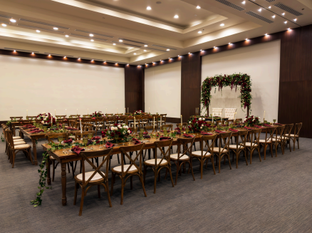 screenshot 20260609 140050 be all inclusive Banquet hall with long wooden tables set for a formal event, with floral centerpieces and a greenery arch in the background